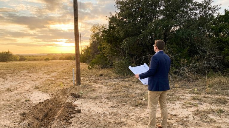 Un homme en costume examinant des plans d'architecture sur un terrain vide au coucher du soleil.